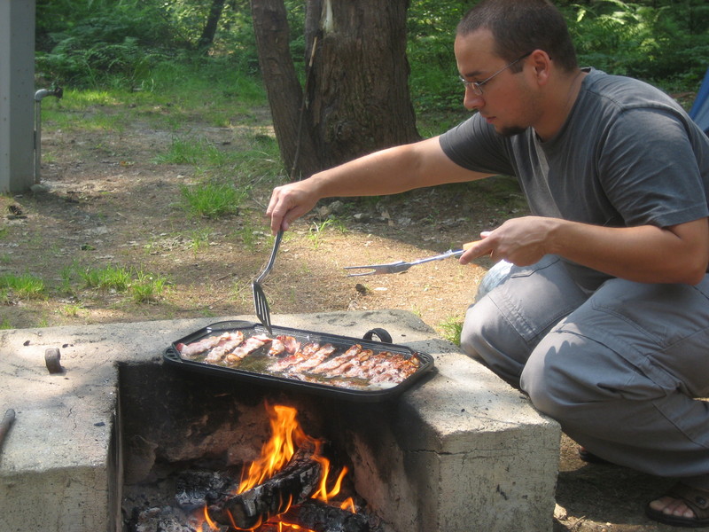 Jay cooking bacon over a camp fire.